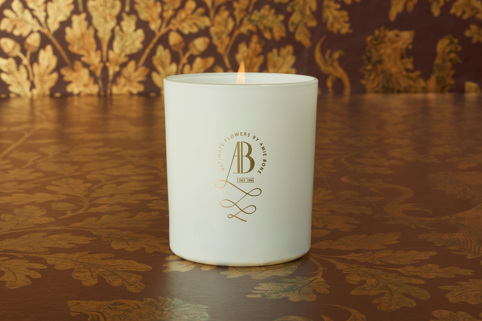 a white candle with an iconic gold crested logo sitting next to a white gift box tied in black ribbon