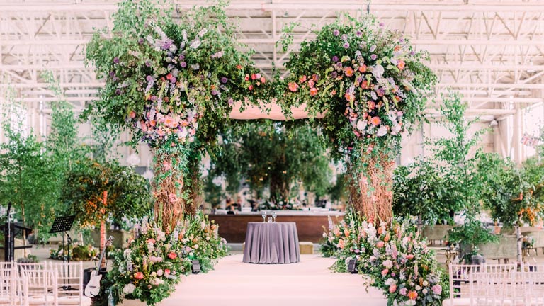 A massive green chuppah bursting with flowers and foliage inside an airport hangar for a luxury Jewish wedding